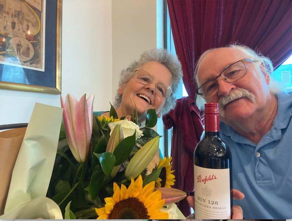2 smiling people with a bunch of flowers and a bottle of wine celebrate their 48th wedding anniversary