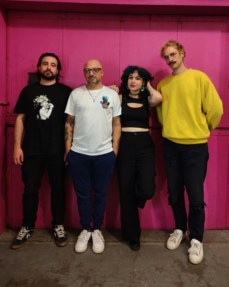 Band of 3 guys and a woman standing in front of a pink wall looking cool and serious 