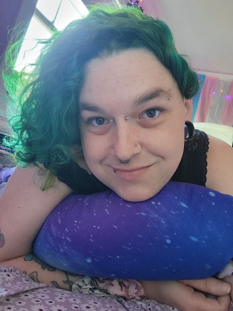 smiling white trans woman with green hair and many piercings, a trans flag apparent in the background 