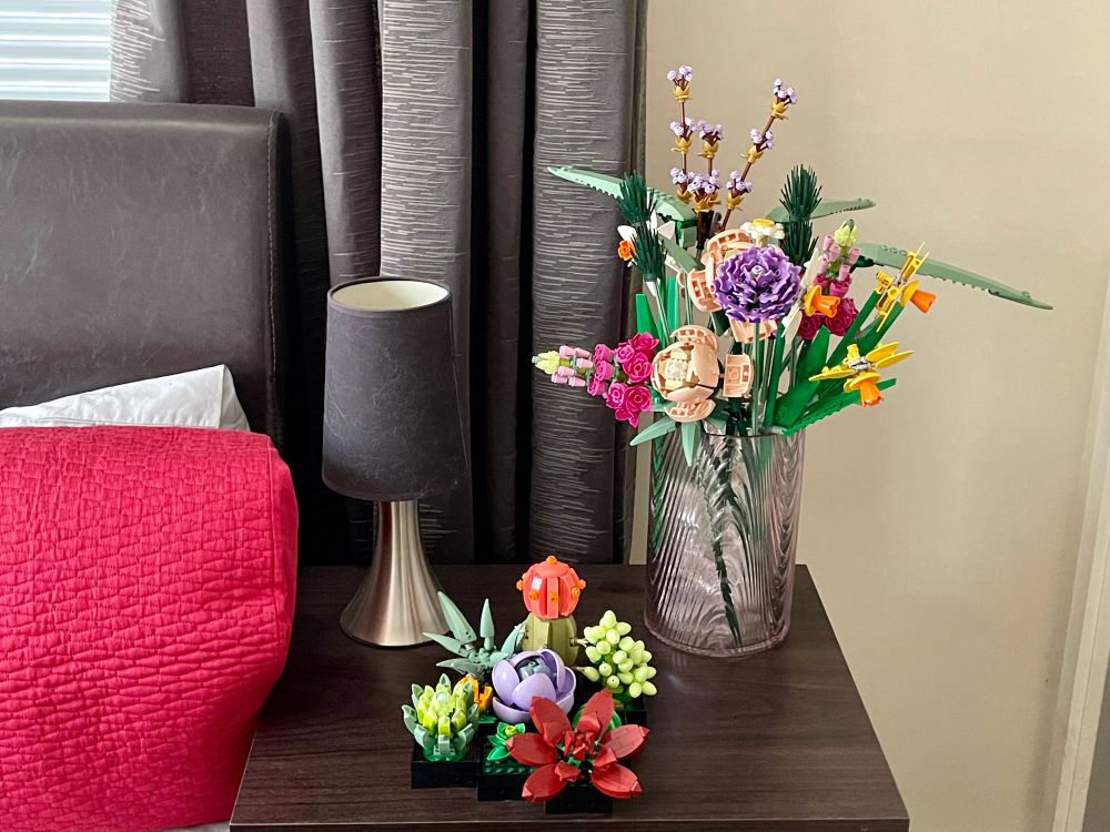 A bedside table with a black and silver lamp, a vase of Lego flowers, and some Lego succulents.