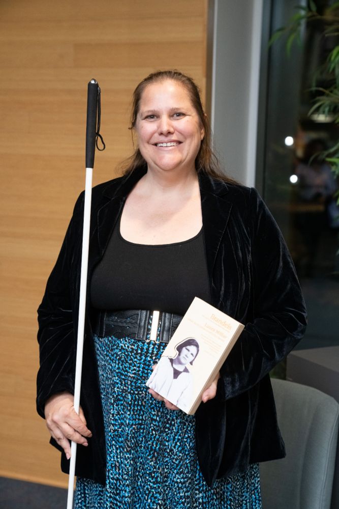 Laura Millar, a brown-haired white woman wearing a black top and blue patterned skirt, smiles in response to a remark.