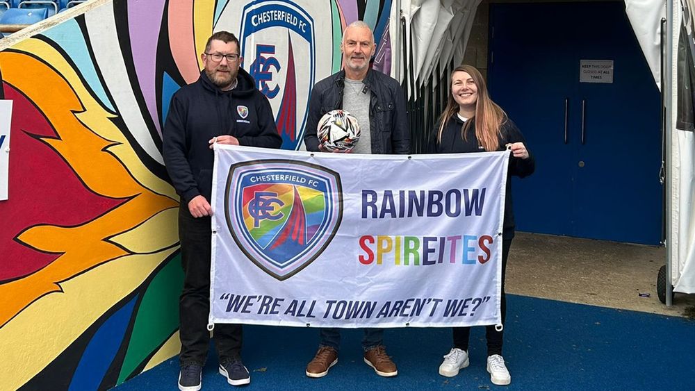 The image shows from left to right, Darren (Co founder of Rainbow Spireites), Jamie (ex Chesterfield FC player) and Ellie (co-founder of Rainbow Spireites, behind the Rainbow Spireites flag which depicts the clubs rainbow badge, 'Rainbow Spireites ' and 'We're All Town Aren't We?' 