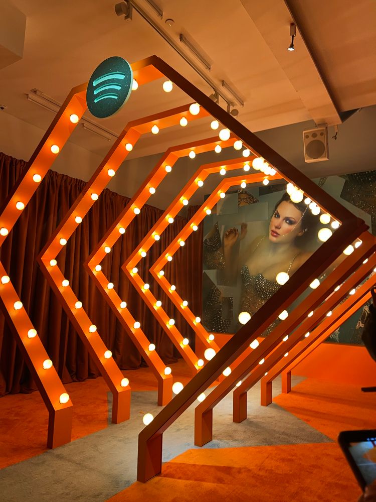 the life of a showgirl album cover on top of the eras tour stage in gray carved into an orange carpet. over the carpet are orange frames with lights all over them. a teal spotify logo is at the top. 