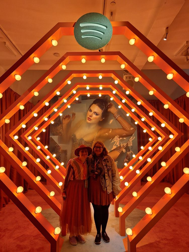 mir and sam pose together in front of the life of a showgirl album cover on top of the eras tour stage carved into an orange carpet. they are surrounded by orange frames with lights all over them. mir is a white non-binary person with orange glasses and no hair wearing an orange, yellow, green, red and pink striped long sleeve shirt, an orange skirt, and an orange hat. sam is a white non-binary person with black glasses wearing a light blue dyed jean jacket over a green taylor swift reputation snake shirt and an orange dress with black tights. 
