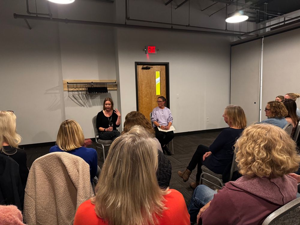 Two authors sit in front of a crowd. The one on the right, in light purple, is looking at the author on the right, wearing black. The backs of multiple women’s heads are seen as the listen to the authors. 