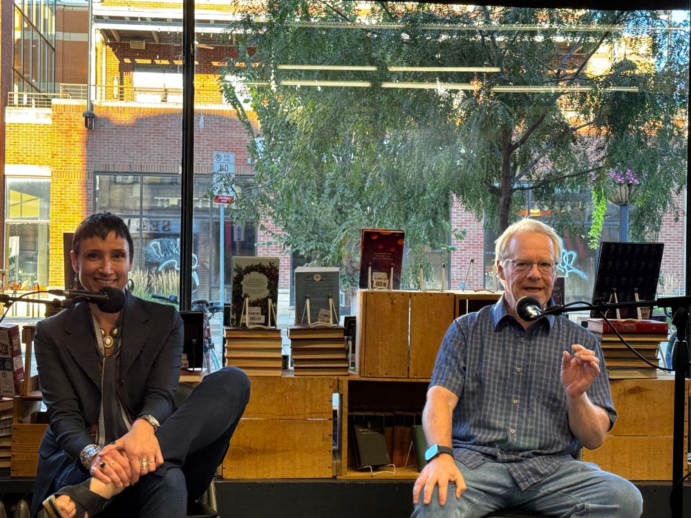 Author Brandy Schillace sits on the left behind a microphone, with legs crossed while smiling. On the right, author Matt Goldman appears to be actively talking. Brandy wears a dark blazer with dark jeans, while Matt wears a blue and white patterned button up shirt with light jeans. There are books in the background and the authors sit in front of a window with a view of trees and buildings outside. 