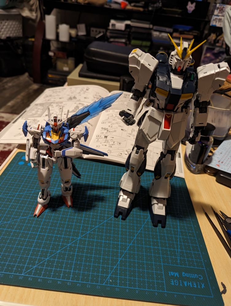 The full body of the Nu Gundam model kit, standing next to an Aerial Gundam model kit on a modeling board