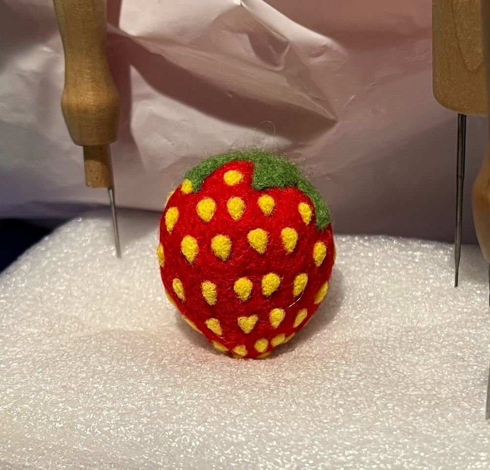 A strawberry needle felted out of wool with one seed in the shape of a heart. Sitting on a foam block. 