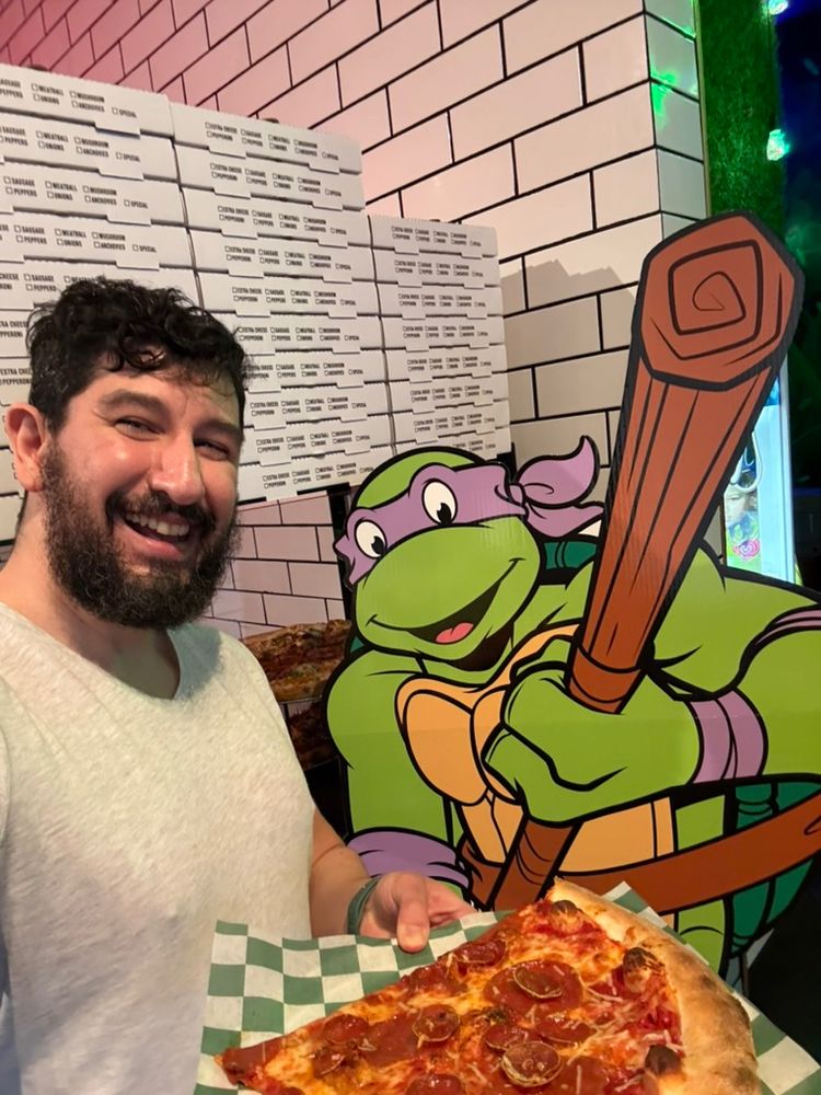 An image of myself holding a big slice of pizza, in front of a poster of Donatello holding his bo staff