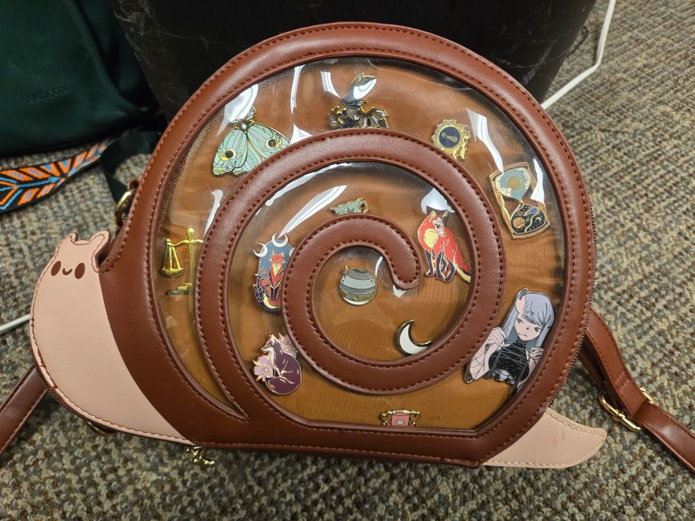 An Ita bag shaped like a snail with an assortment of anime and pop culture pins.