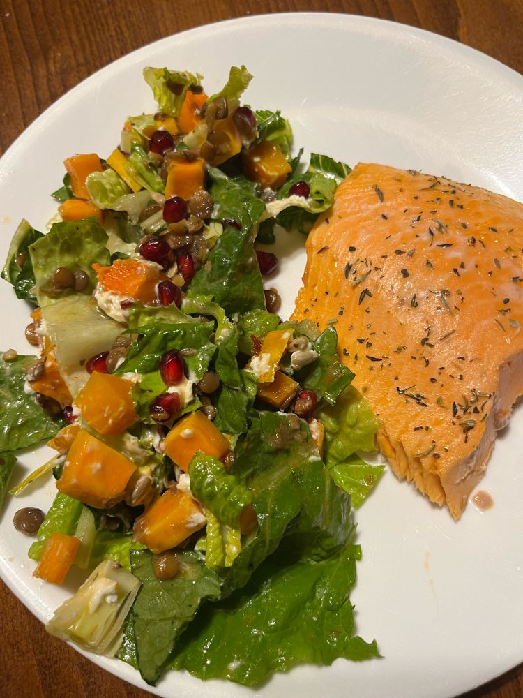 Roasted salmon on a white plate with a salad of romaine, pomegranate seeds, chèvre, lentils, sunflower seeds, & roasted butternut squash 