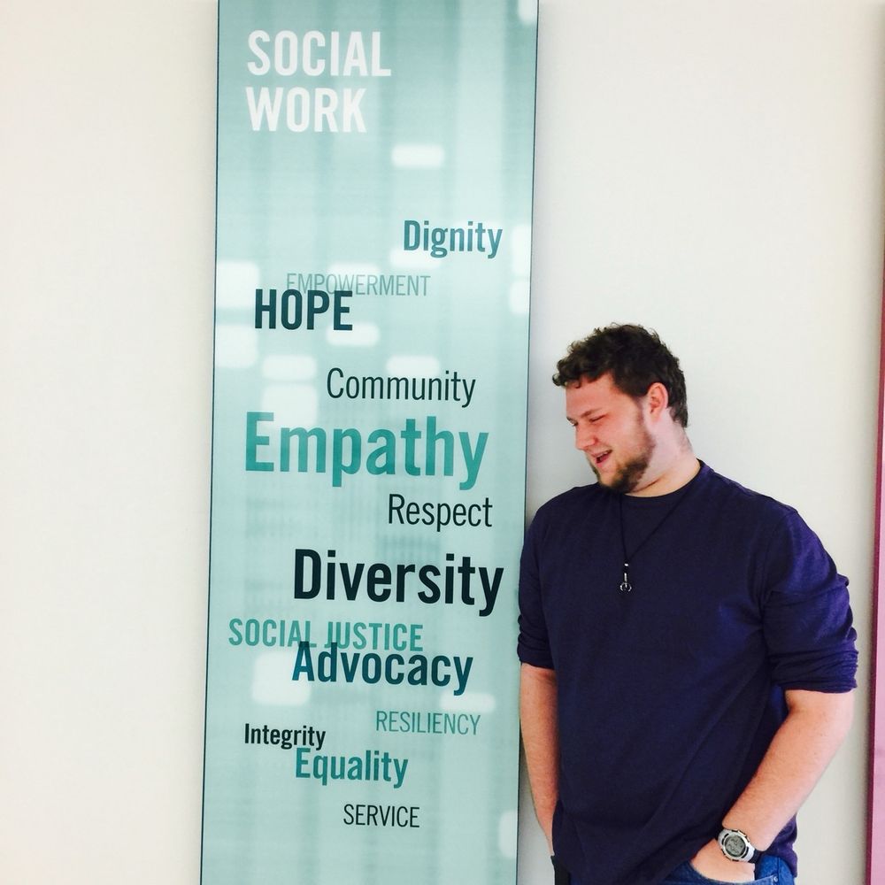 A white masculine person in a purple shirt with their sleeves rolled up and short brown hair looks at a teal sign that says, “Social Work, Dignity, Empowerment, Hope, Community,  Empathy, Respect, Diversity, Social Justice, Advocacy, Resiliency, Integrity, Equality, & Service.”
