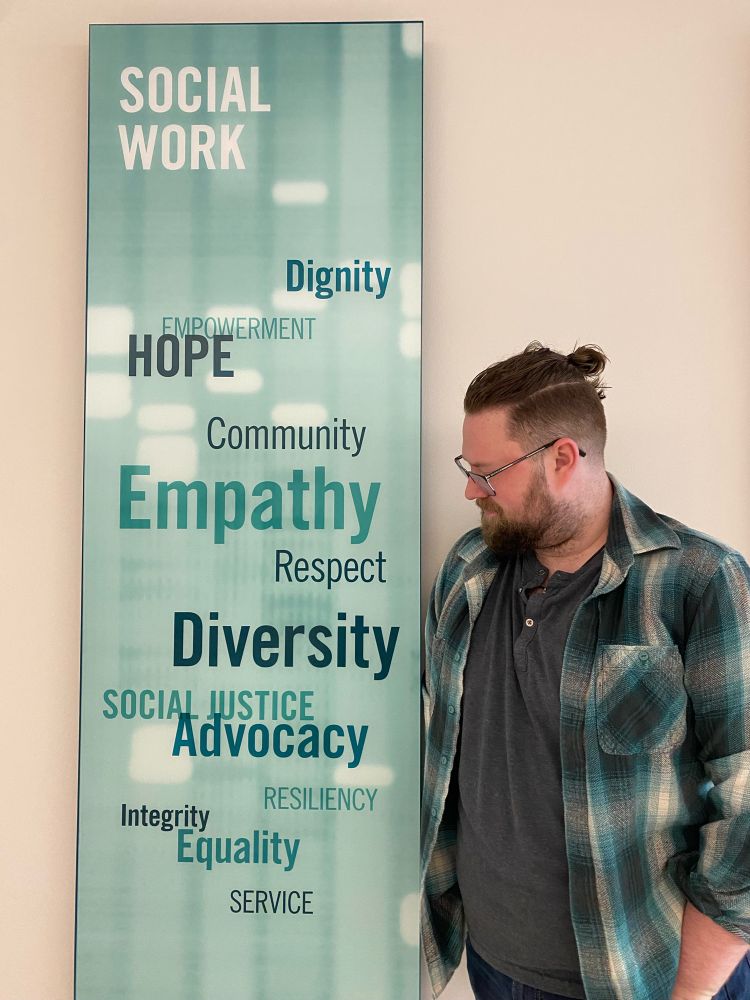 A white masculine person in a teal flannel and grey undershirt with their hair in a bun looks at a teal sign that says, “Social Work, Dignity, Empowerment, Hope, Community,  Empathy, Respect, Diversity, Social Justice, Advocacy, Resiliency, Integrity, Equality, & Service.”
