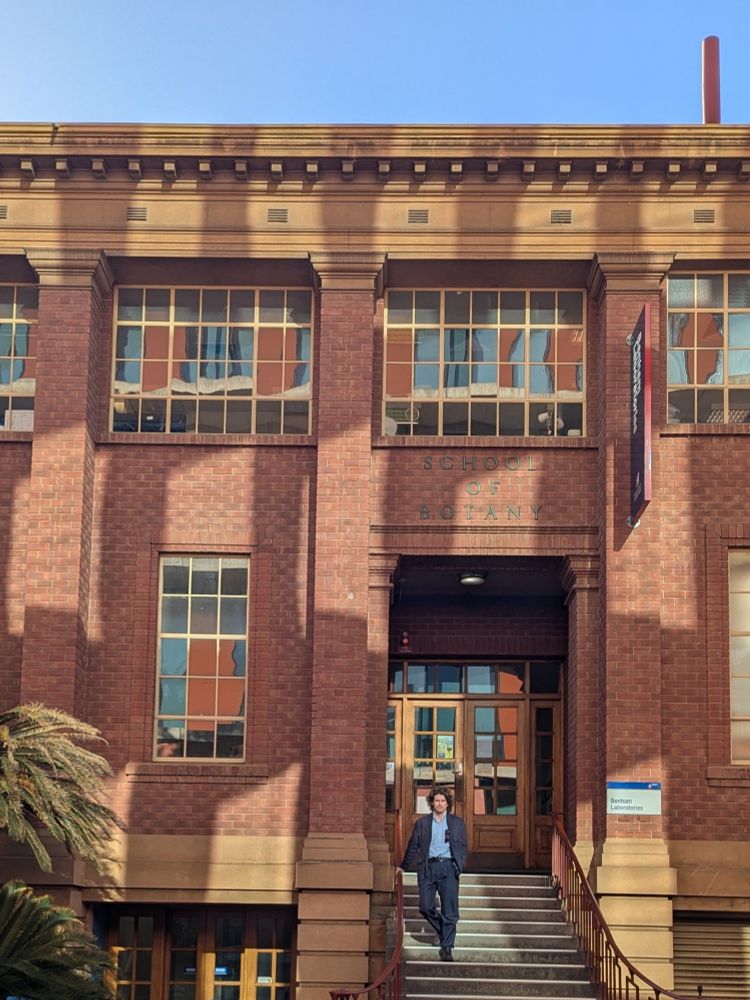 Three storey red brick and sandstone building with sign "school of botany" with Joe standing on steps in main entrance 