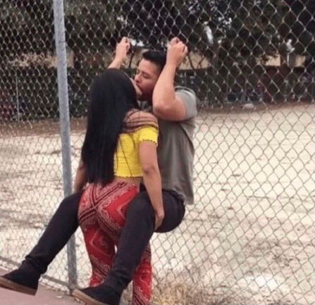 Woman holding a man up by his legs as they kiss and he’s holding onto the chainlink fence behind him