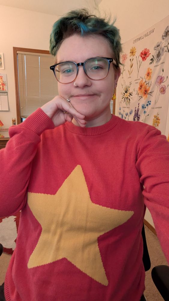Selfie of me wearing a red sweater with a big yellow star on it. I have short greenish blue hair and I'm sweating glasses and small rubber duck earrings