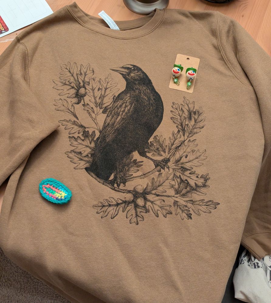 A brown sweater with black are of a crow on branches with acorns on it. On top of the sweater is a pair of green clown earrings, and a colorful crochet fidget toy 