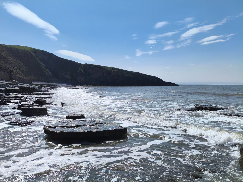 Looking east in Dunraven Bay. There are flat, wave wetted, rocks leading off through the surf towards a dark cliff that slopes down to the sea. The sun is high, out of left of frame, giving strong shadow accounting for the dark cliff.