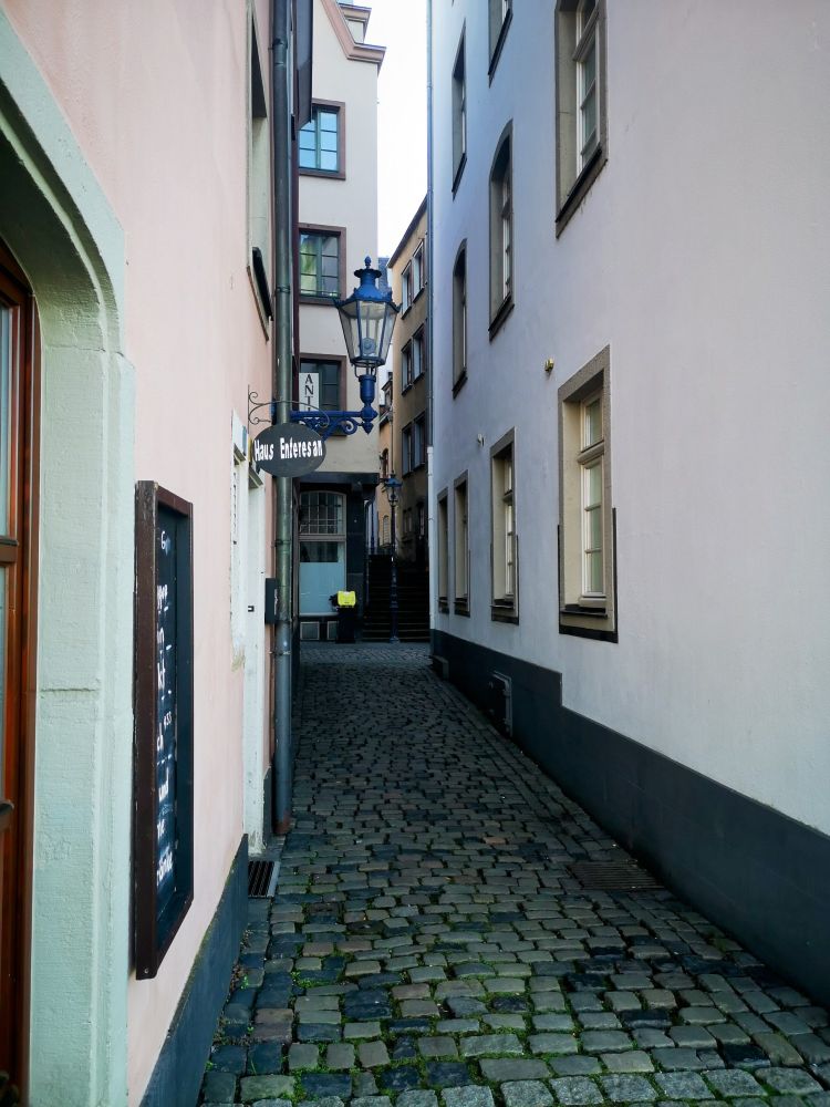 A narrow back street in Köln, leading away from the Rhine. White walls, Granite sets, a blue victorian era street lamp on a wall bracket. The vanishing point is surrounded by a yellow topped Wheelie bin, a gas light style standard streetlight and a flight of steps leading g to infinity.