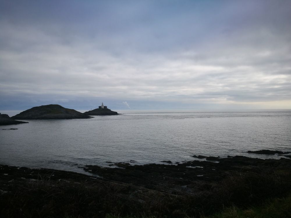 Sea scape of Swansea Bay. Rocky foreground, mumbles lighthouse centre left and the plume from Port Talbot steelwork in the distance. Grey,  calm, sea and and flat grey clouds. 