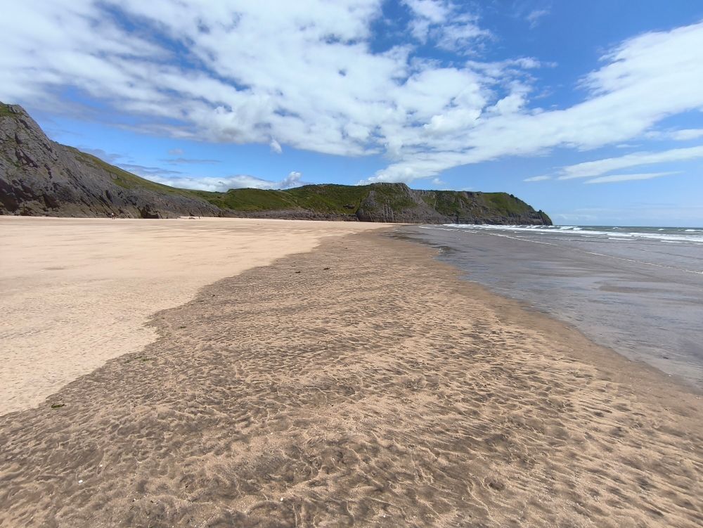 A sandy beach, cliffs to the left, sea to the right. The view point is at the point at which the incoming waves sink into the sand on a rising tide. Breaking waves are seen in the far right of the pucture. The sky is blue with scattered clouds.