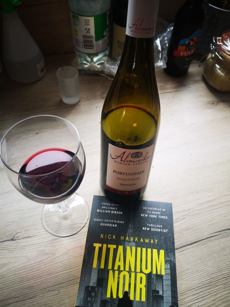 An opened bottle of Ahrweiler Portugieser red wine, a glass of the same and a copy of Titanium Noir by @harkaway.bsky.social arranged to illustrate the new best way to spend time on Christmas Day. 