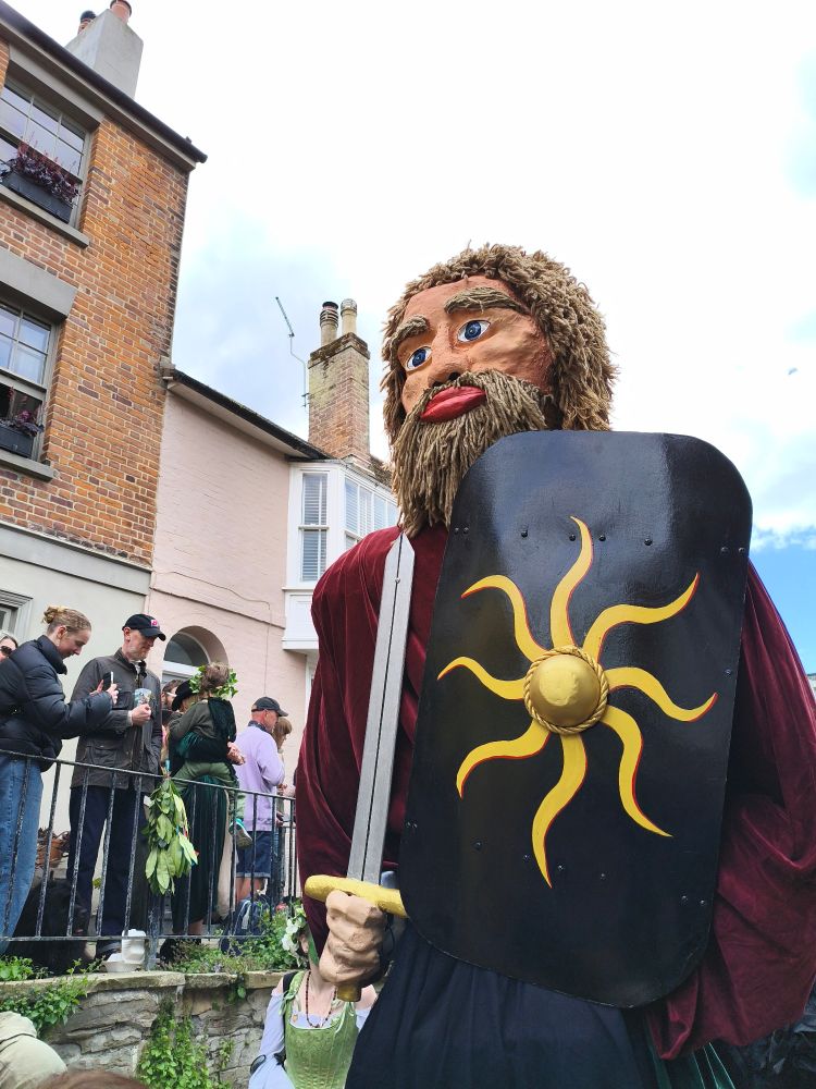 A giant walks down Hastings Old Town High street. He has a gladius in his right hand and a black shield decorated with a sun figure in his left.