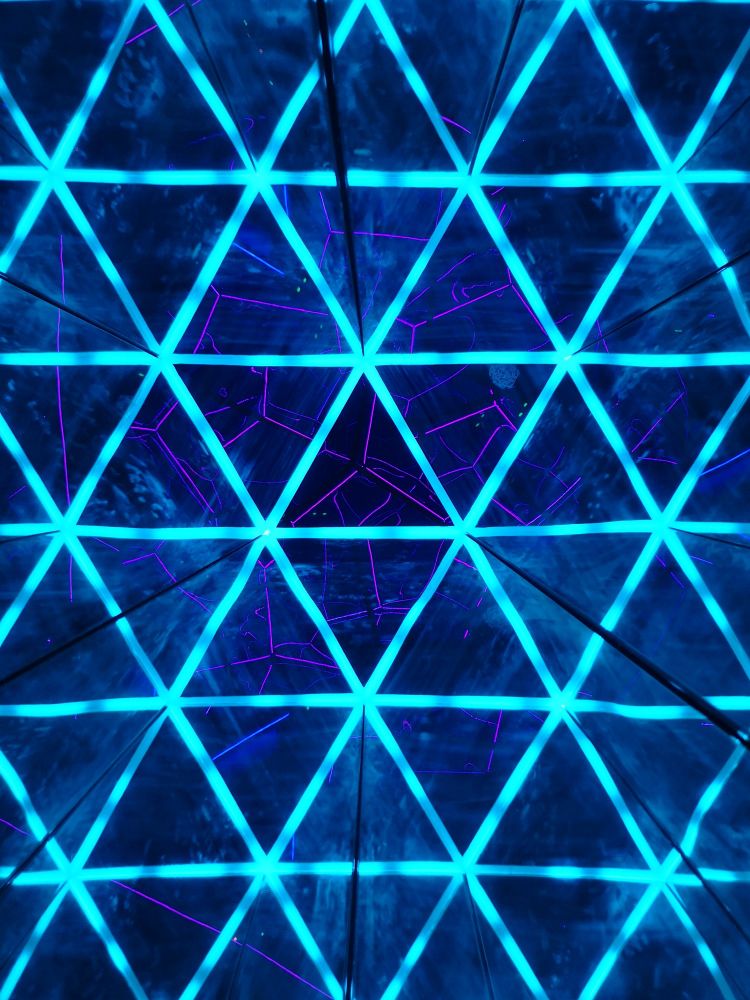 A mesh of lit cyan triangles in front of a magenta tracery. Formed by taking a picture down the inside of an illuminated, mirrored triangular prism pointed at an illuminated, transparent, dodecahedron. 