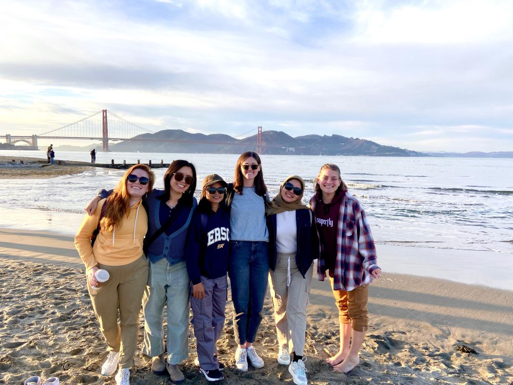 Six women stand smiling on a beach in front of the Golden Gate Bridge. 