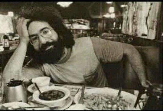 Jerry Garcia in a diner booth leans, head on hand, to his side, looking at the camera. The table ahead of him is full of plated food, a bowl of soup, and coffee.
