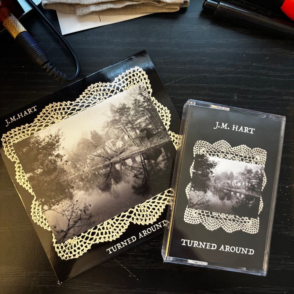Cassette and cd editions of J.M. Hart’s forthcoming album, “Turned Around”. On each, the background is black with the artist and album names respectively above and below a doily with a black & white photograph of a river.

These all sit on a black tabletop. A headphone wire is seen plugged in at the upper left. The edge of a folded handkerchief is at the top.