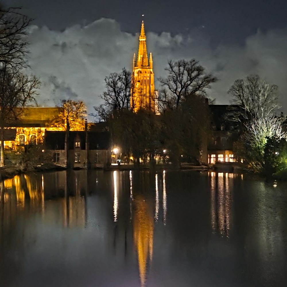A floodlit church tower with white clouds & dark night sky behind. The church tower is reflected in water in the lake in foreground.