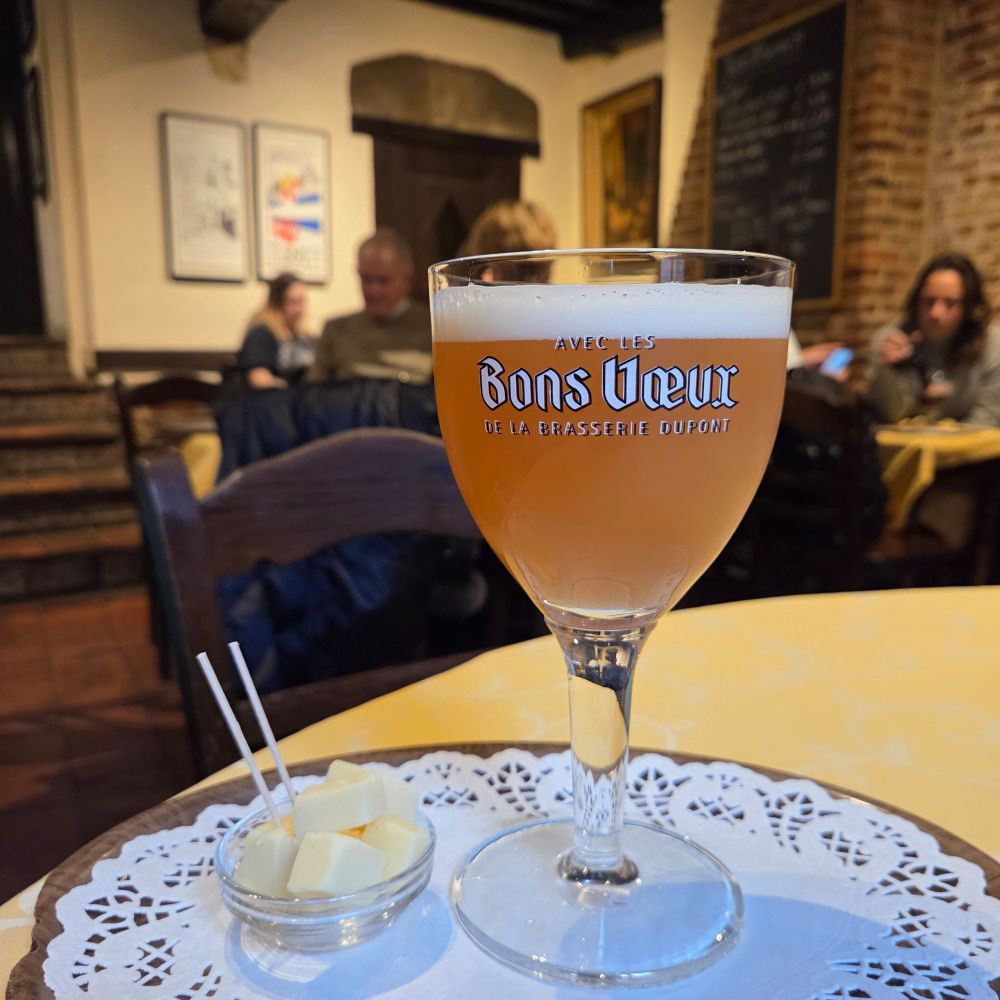 A glass of draft Avec Les Bons Vœux beside a small dish of cheese.