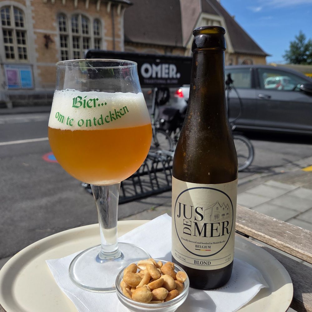 A blond beer in its glass with bottle to right. There's a small dish of nuts in between. 