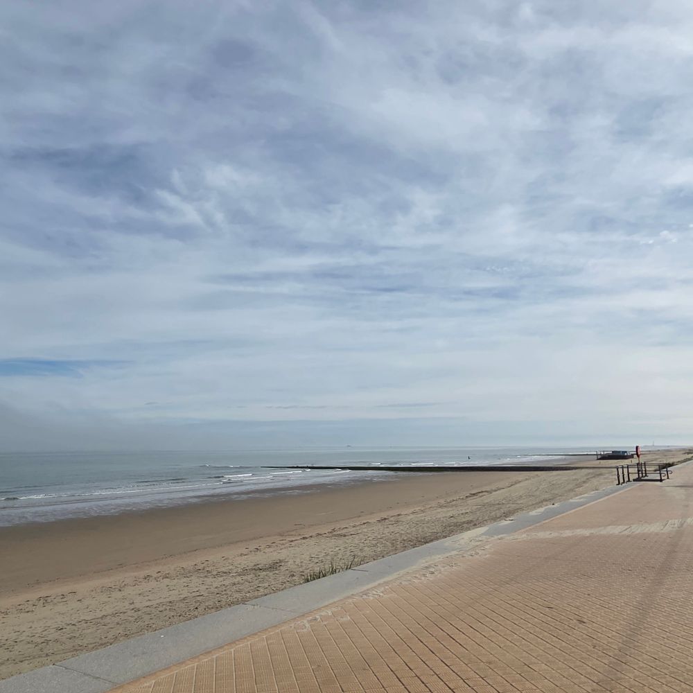 A sandy beach with path to the right. Sea is to the left. Skies above are slightly cloudy and pale blue.