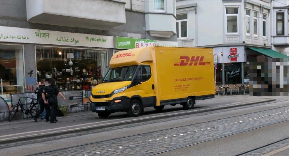 Zwei Mitarbeitys des Ordnungsamts, die auf einem an einer Straßenbahnhaltestelle und auf einem Blindenleitsystem geparkten Lieferwagen zulaufen. Sie laufen daran vorbei, ohne etwas zu unternehmen.