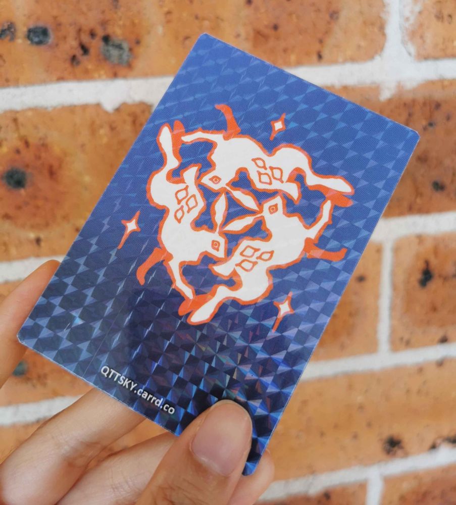 A photo of the backside of the playing card. It is blue and holographic, with a symbol comprised of 3 hares prancing in a circle, ears intertwined.