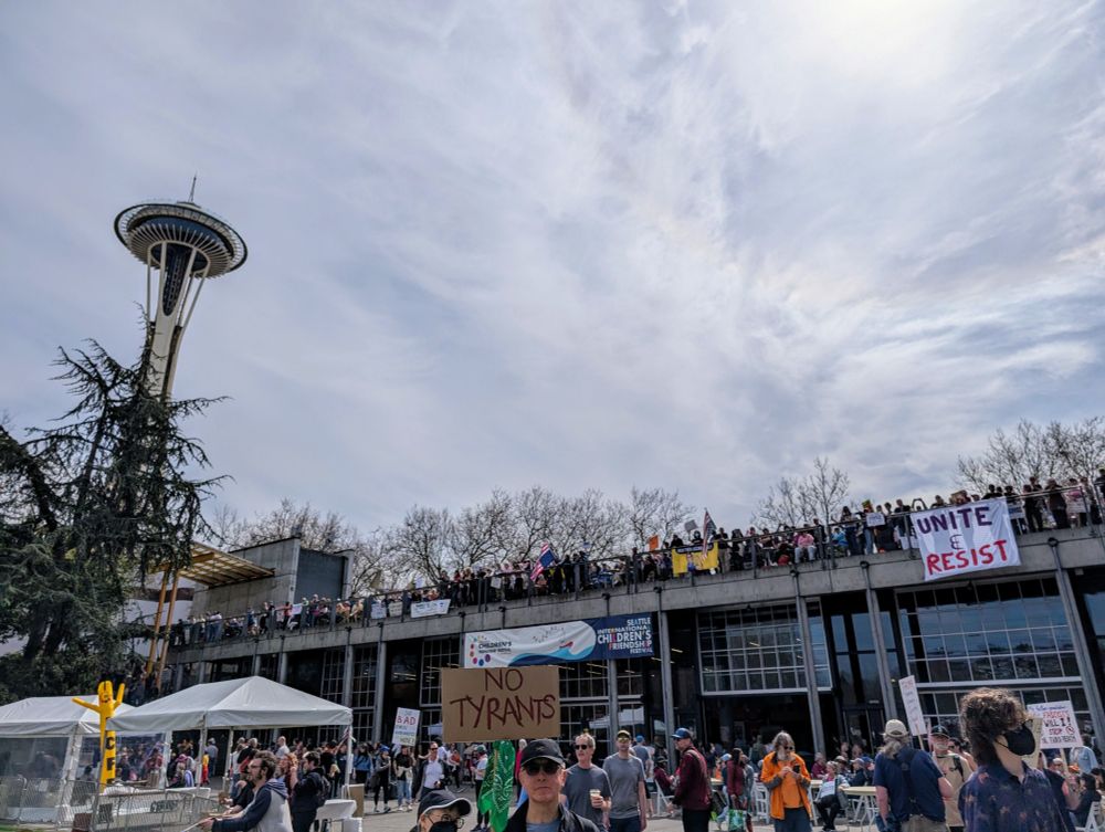 Many people out at a rally. A sign saying "unite and resist" hangs off a balcony and the space needle is visible in the background 