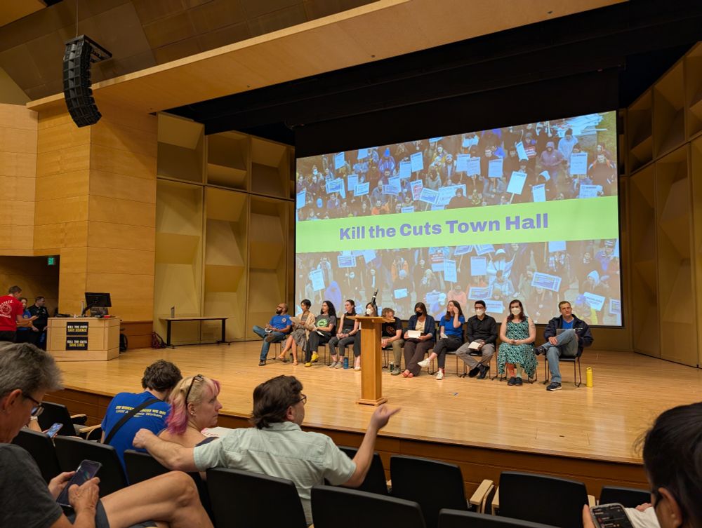 A stage with a panel of people sitting on it. A projector is displaying a picture of protestors and the slogan "kill the cuts" behind the panel.