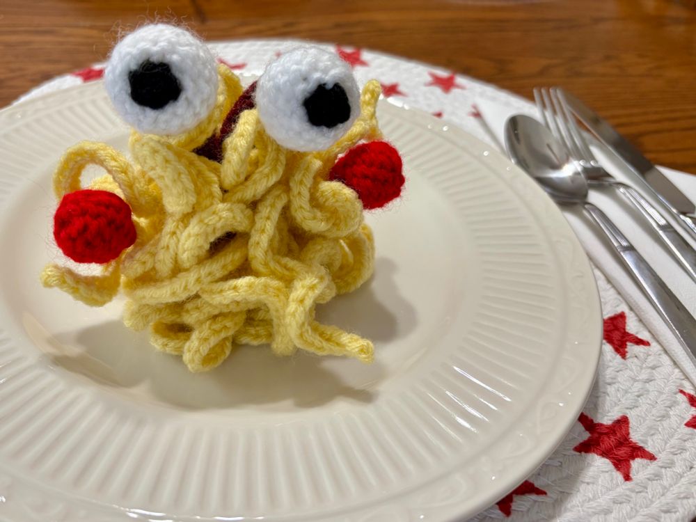 A slip stitch crochet spaghetti monster finger puppet, served on a white ceramic plate. It’s a mass of noodly light yellow tentacles, with two red meatballs, and eyeballs on maroon stalks.