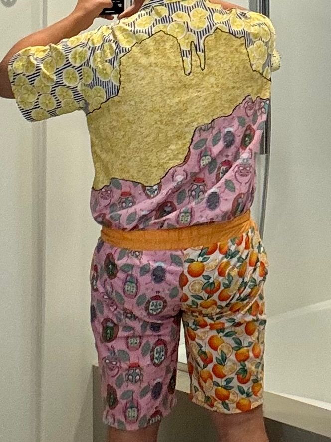 Rear view of a terrifying outfit, comprised of a shirt and shorts in a loud and senseless combination of patterns and colors. Bright oranges and yellows, a stripey lemon pattern, a pink fabric with fanciful beetles, and a fabric with oranges scattered everywhere. The shirt features a satin stitch appliqué technique, meant to look like an abstract mountainous landscape. The shorts are wrinkled and puckered around the but, due to poor easing in the crotch.