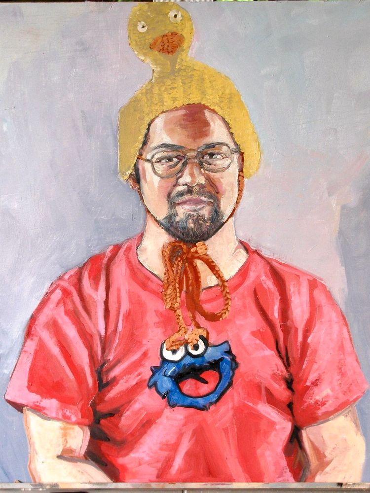 Portrait of a goofy-looking man with a serious expression, wearing a crocheted duck hat, glasses, and a red t-shirt featuring Cookie Monster’s face. Neutral, slightly lavender background.