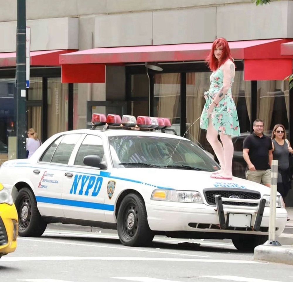 A red haired babe in a sundress with a delicate floral pattern, and flip flops standing on top of an NYPD cop car and pissing with a pretty impressive stream.