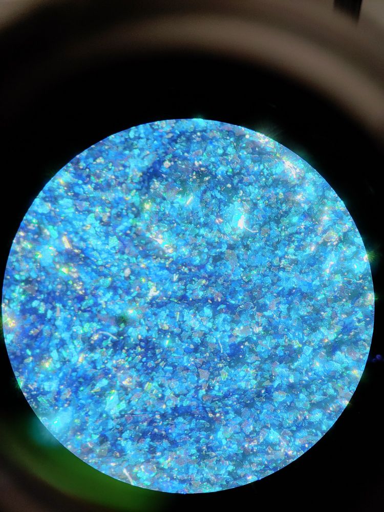 View through microscope with a circular perspective showing blue glittering shiny crystal-like structures in several different shades. The surface structure looks a bit  like unpolished granit. A few greenish and yellow accents are visible.