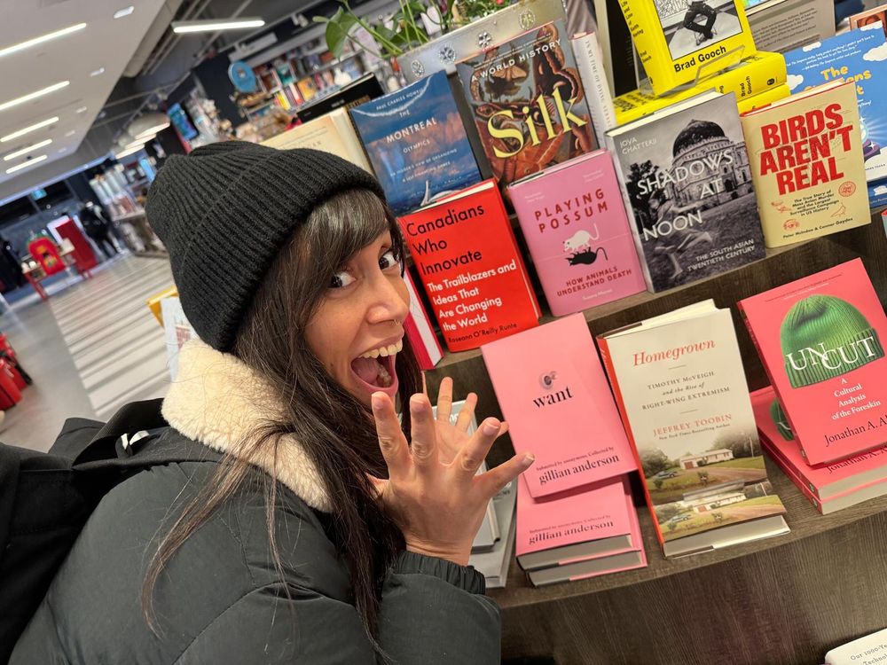 A woman with dark hair wearing a black puffer and beanie looking excited in front of a table full of books. In the centre is a copy of Playing Possum, the book she’s excited about.