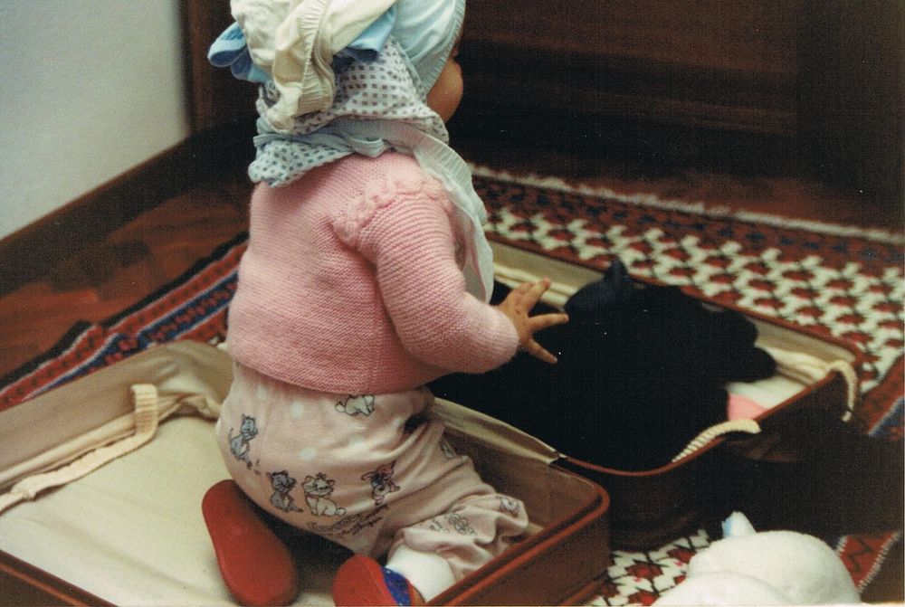 A toddler sitting inside a brown suitcase, wearing a pink Aristocats pijama and a heap of underpants on her head.