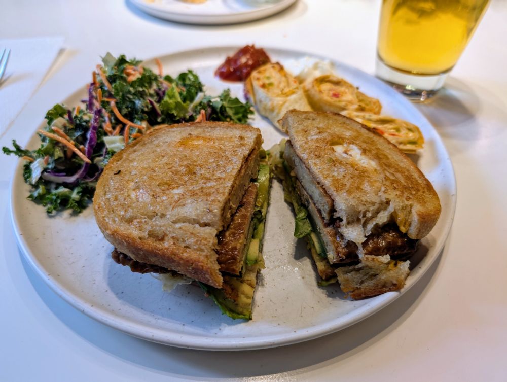 A white plate holding a tempeh Reuben with avocado and sauerkraut,  green salad and a few antojitos on the side