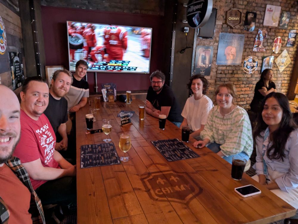 A group of eight people sitting around a long wooden table in a pub, smiling at the camera during the UmbraCymru summer social. Drinks are on the table, and a large screen in the background shows a Formula 1 pit lane scene. The venue has exposed brick walls decorated with beer signs and posters, creating a relaxed and friendly atmosphere.