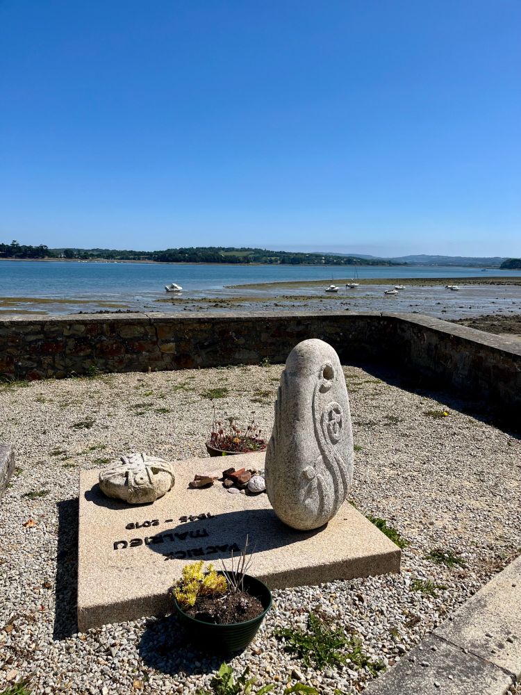 Picture of a Celtic tombstone standing in front of the shoreline. It is low tide and boats are laying on their side. The stone has the symbol of the "Hermine" also represented on the Breton flag.  