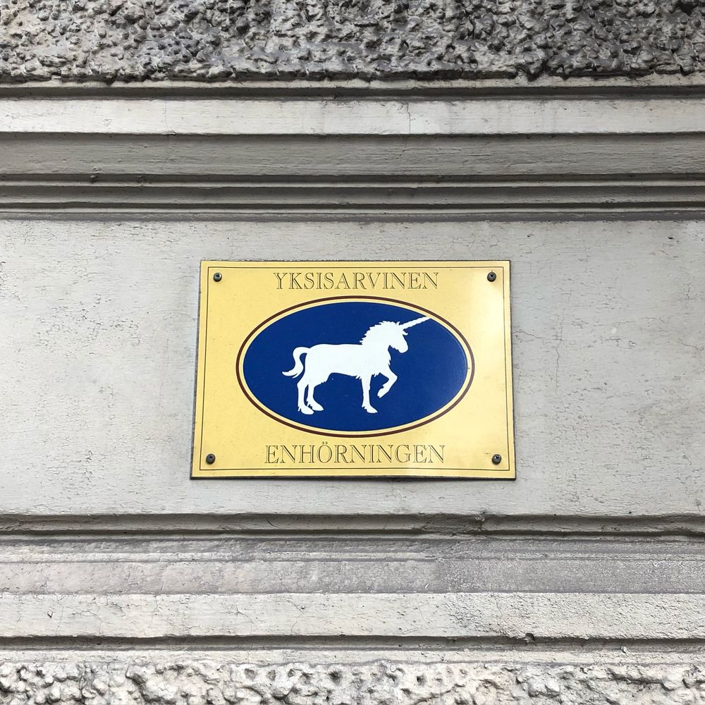 Photo shows a street sign in Helsinki. A yellow gold rectangle has text that reads ‘YKSISARVINEN ENHÖRNINGEN’ and in the centre is a blue oval with a white unicorn figure on it.